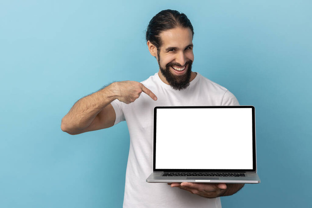 Porträt eines Mannes mit Bart, der ein weißes T-Shirt trägt, einen Laptop mit leerem Bildschirm hält und in die Kamera lächelt, Internetwerbung. Indoor Studio aufgenommen isoliert auf blauem Hintergrund. - Foto, Bild