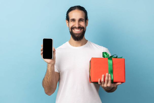 Present, bonus for mobile user. Handsome happy man with beard holding gift box and cell phone with mock up, blank display for advertise. Indoor studio shot isolated on blue background. - Zdjęcie, obraz