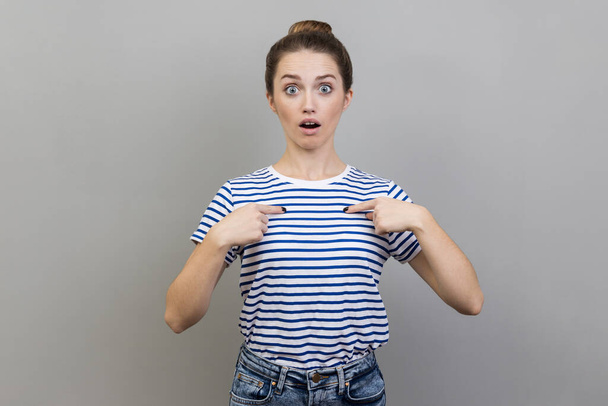 Portrait of astonished woman wearing striped T-shirt pointing at herself, asks who me, has surprised expression, shocked being picked. Indoor studio shot isolated on gray background. - Fotoğraf, Görsel