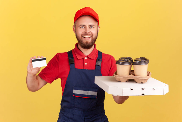 Portrait of smiling delivery man standing with pizza box and coffee in disposable cup and showing credit card, online paying for delivery. Indoor studio shot isolated on yellow background - Foto, afbeelding