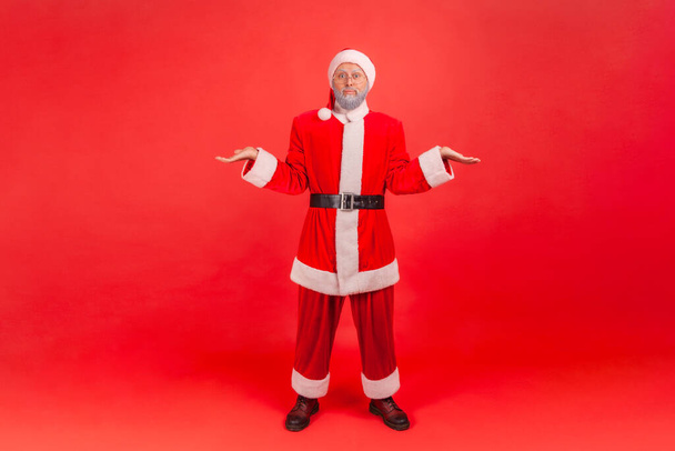 Full length portrait of elderly man with gray beard wearing santa claus costume shrugging shoulders as doesn't know answer, being uncertain, not sure. Indoor studio shot isolated on red background. - Photo, Image