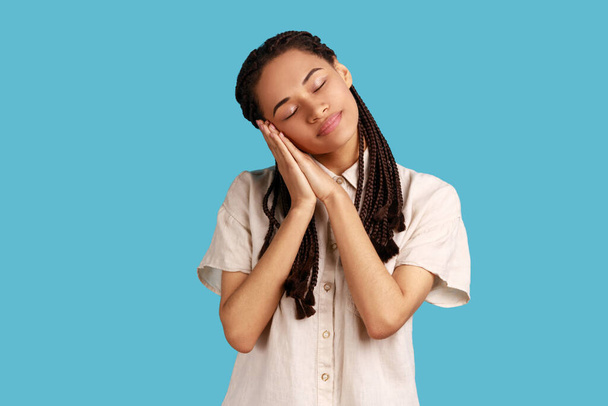 Portrait of beautiful woman with black dreadlocks sleeping laying down on her palms, having comfortable nap and resting, dozing off, wearing white shirt. Indoor studio shot isolated on blue background - 写真・画像