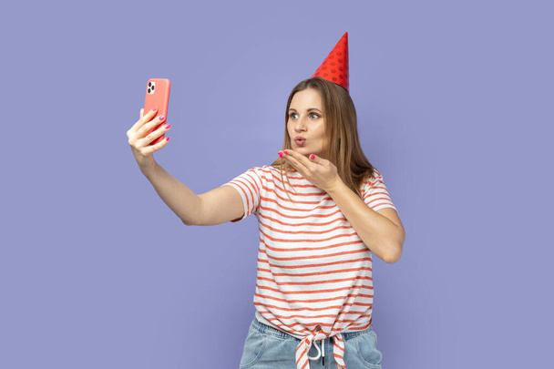 Portrait of happy blond woman wearing striped T-shirt and party cone, having video call or broadcasting livestream, sending air kisses. Indoor studio shot isolated on purple background. - Zdjęcie, obraz