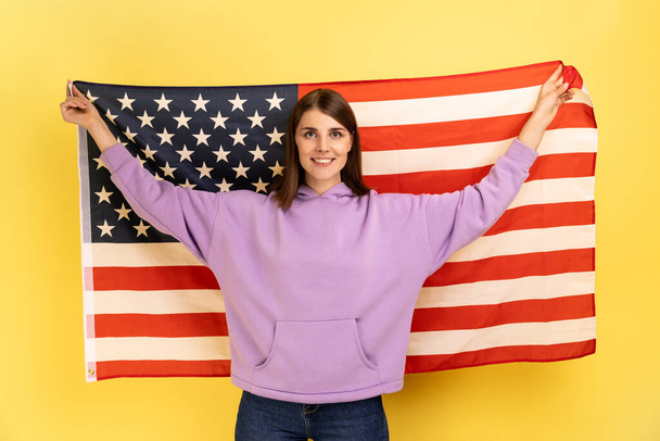 Portrait of attractive happy positive woman standing with raised arms, holding USA flag, celebrating national holiday, wearing purple hoodie. Indoor studio shot isolated on yellow background. - Photo, image