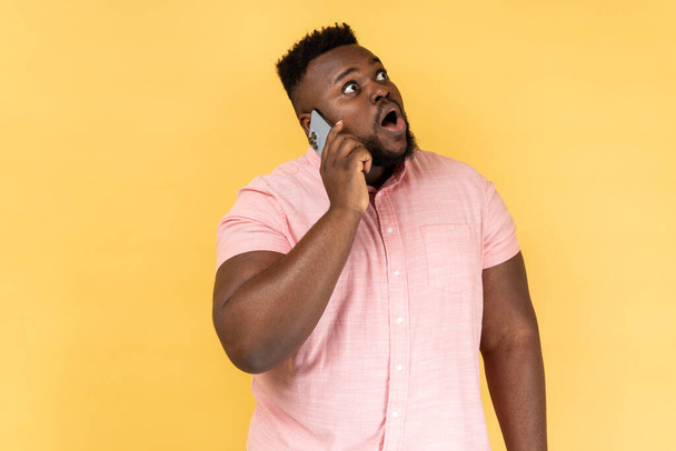 Portrait of shocked man wearing pink shirt talking with friend on cell phone, looking away with surprised amazed, hearing breaking news. Indoor studio shot isolated on yellow background. - Foto, Bild