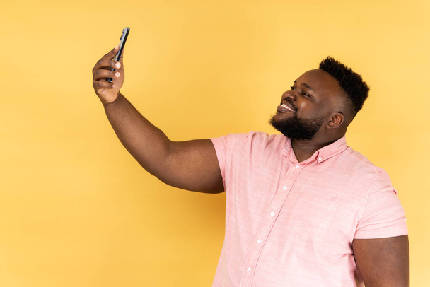 Portrait of positive optimistic man wearing pink shirt talking on video call, having online conversation on mobile phone, taking selfie. Indoor studio shot isolated on yellow background. - Foto, afbeelding