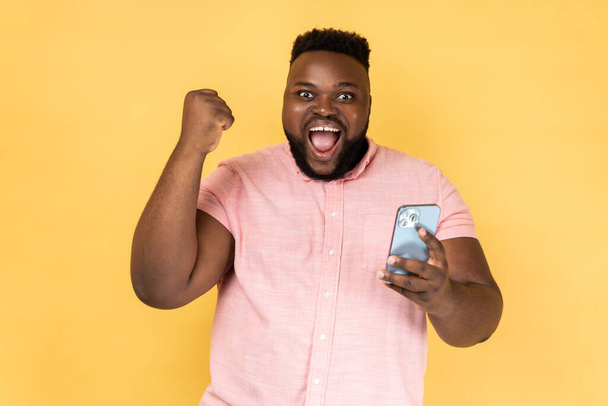 Portrait of satisfied bearded man wearing pink shirt standing with cell phone in hands and clenching fist, celebrating his victory, looks at camera. Indoor studio shot isolated on yellow background. - Fotografie, Obrázek
