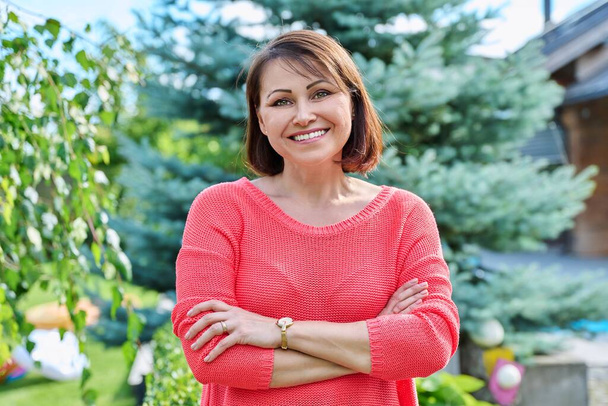 Portrait of smiling middle aged woman looking at camera outdoor. Successful confident mature woman in red with arms crossed, nature green lawn background - Fotoğraf, Görsel