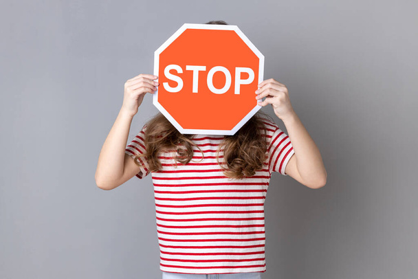 Little girl in striped T-shirt covering face with Stop symbol, anonymous woman holding red traffic sign stop, warning of danger, restriction and limits. Indoor studio shot isolated on gray background. - Photo, Image