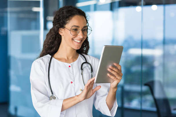 Latin American doctor woman using tablet computer, female doctor working inside office building wearing white medical coat. - Photo, Image