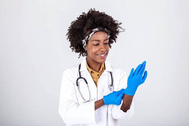 Retrato de una atractiva joven doctora en bata blanca poniéndose guantes médicos. Retrato de médico sonriente alegre en uniforme blanco de pie con las manos cruzadas sobre fondo gris - Foto, imagen