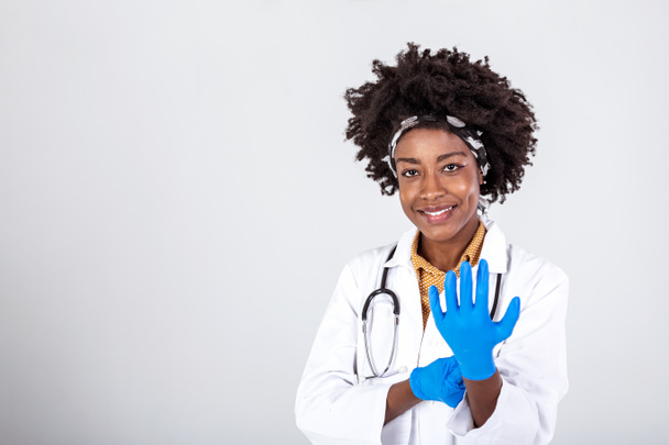 Porträt einer attraktiven jungen Ärztin in weißem Mantel, die medizinische Handschuhe anzieht. Porträt eines glücklich lächelnden Arztes in weißer Uniform, der mit verschränkten Händen vor grauem Hintergrund steht - Foto, Bild