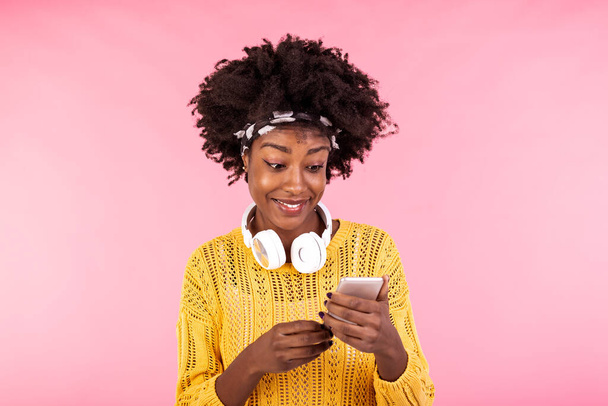 Afrikai amerikai nő fülhallgatóval és mosollyal. Fiatal nő természetes göndör haj gazdaság és nézi a mobiltelefon. - Fotó, kép
