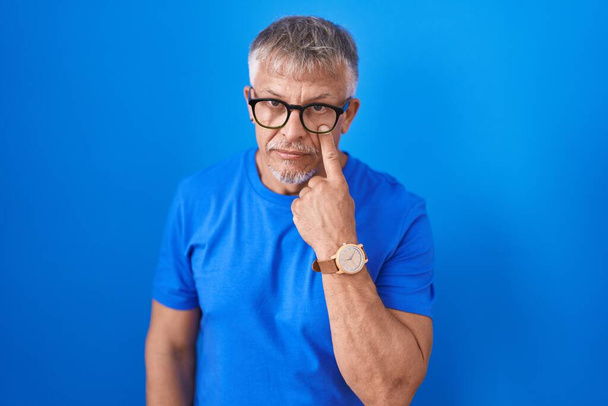 Homem hispânico com cabelos grisalhos em pé sobre fundo azul apontando para o olho observando você gesto, expressão suspeita  - Foto, Imagem