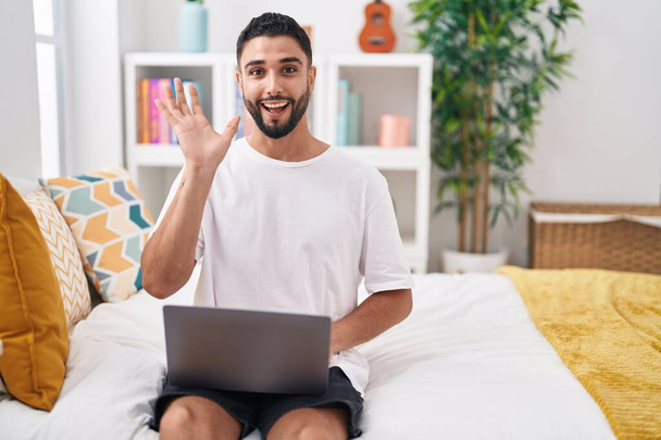 Ισπανόφωνος νεαρός άνδρας που χρησιμοποιεί φορητό υπολογιστή κάθεται στο κρεβάτι γιορτάζει τη νίκη με χαρούμενο χαμόγελο και νικητής έκφραση με υψωμένα χέρια  - Φωτογραφία, εικόνα