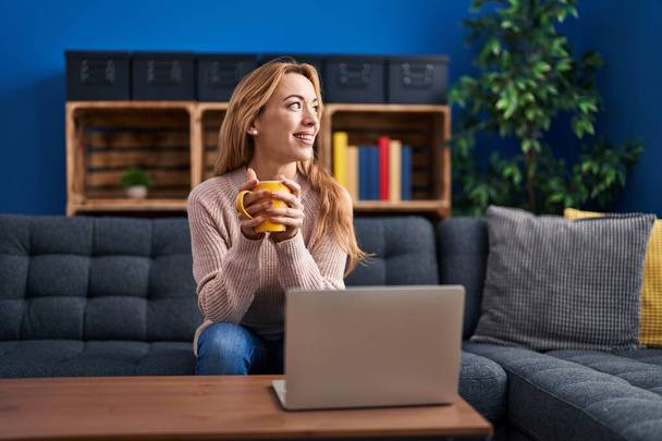 若いです女性を使用してノートパソコンとコーヒーを飲みますソファに自宅で - 写真・画像