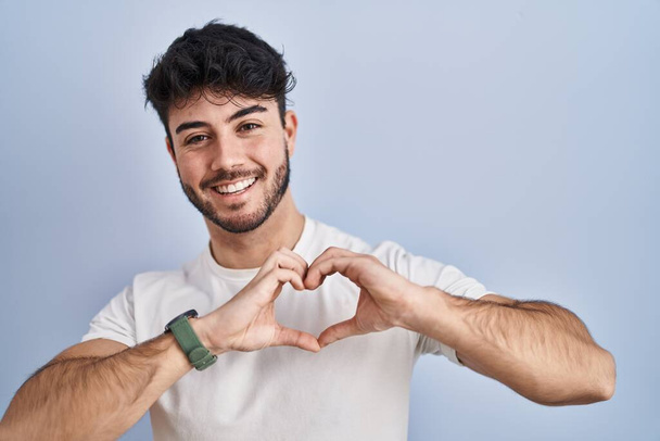 Hispanischer Mann mit Bart, der über weißem Hintergrund steht und verliebt lächelt und Herzsymbolform mit den Händen ausübt. Romantisches Konzept.  - Foto, Bild