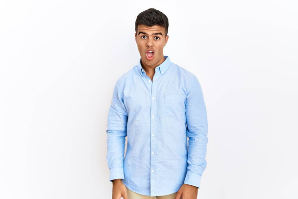 Young hispanic man wearing business shirt standing over isolated background in shock face, looking skeptical and sarcastic, surprised with open mouth  - Фото, изображение