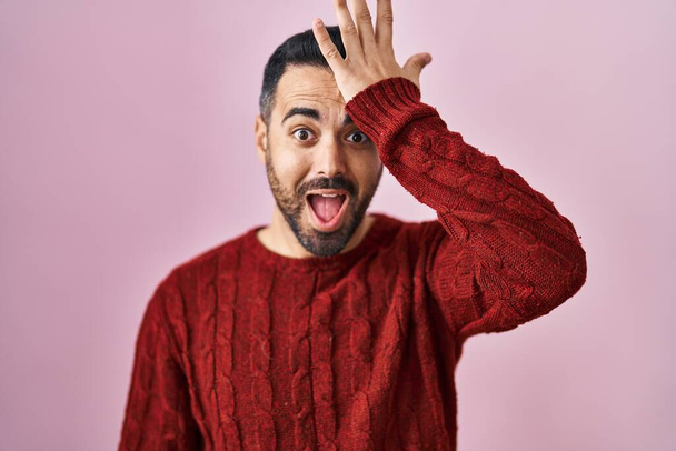 Young hispanic man with beard wearing casual sweater over pink background surprised with hand on head for mistake, remember error. forgot, bad memory concept.  - Foto, Imagen