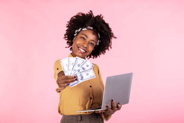 ノートパソコンを使ってピンクの背景に隔絶された明るいアフリカ系アメリカ人女性が紙幣を持っている姿。ノートパソコンを持っている笑顔の女の子の肖像画 - 写真・画像