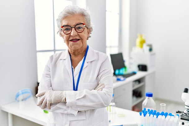 Mujer mayor de cabello gris vistiendo uniforme científico de pie con los brazos cruzados gesto en el laboratorio - Foto, imagen
