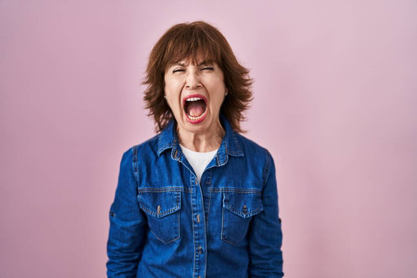 Középkorú nő áll a rózsaszín háttér dühös és őrült sikoltozó frusztrált és dühös, kiabál a harag. düh és agresszív koncepció.  - Fotó, kép