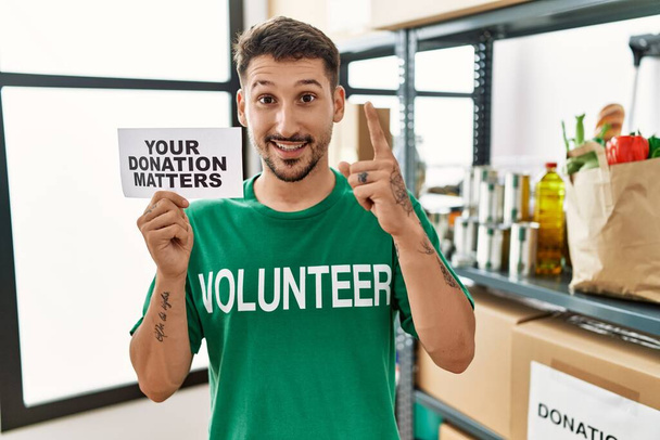 Il giovane volontario che tiene il tuo banner sulle donazioni è sorpreso da un'idea o da una domanda che punta il dito con la faccia felice, numero uno.  - Foto, immagini