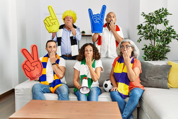 Grupo de personas mayores que apoyan al equipo de fútbol en casa cubriendo la boca con la mano, conmocionados y temerosos de equivocarse. expresión sorprendida  - Foto, imagen