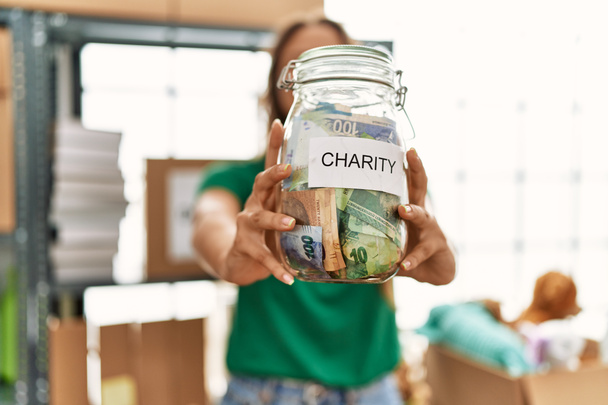 Jovem bela mulher hispânica voluntária segurando frasco dinheiro caridade no centro de caridade - Foto, Imagem