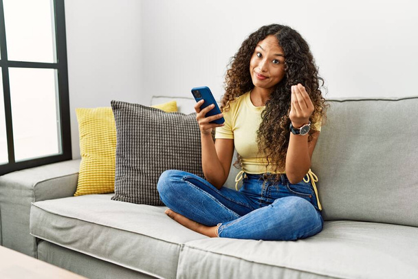 Piękna latynoska kobieta siedząca na kanapie w domu za pomocą smartfona wykonując gesty pieniężne rękami, prosząc o wypłatę pensji, biznes milionera  - Zdjęcie, obraz