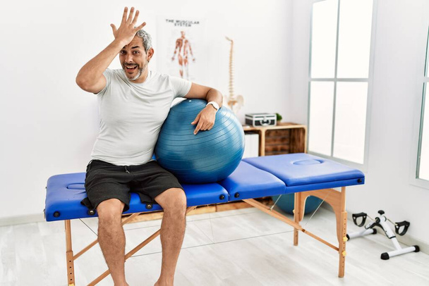 Middle age hispanic man at pain recovery clinic holding pilates ball surprised with hand on head for mistake, remember error. forgot, bad memory concept.  - Photo, Image