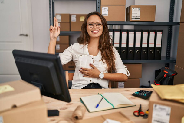 Junge hispanische Frau, die in einem kleinen E-Commerce-Geschäft arbeitet, lächelt und flucht mit der Hand auf Brust und erhobenen Fingern und leistet einen Treueschwur  - Foto, Bild