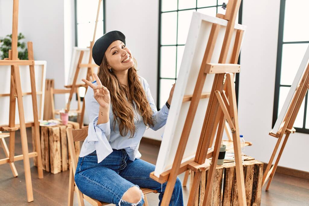 Mladá hispánská umělkyně maluje na plátno v ateliéru a usmívá se s veselou tváří, mrká prsty na kameru a dělá znamení vítězství. číslo dvě.  - Fotografie, Obrázek