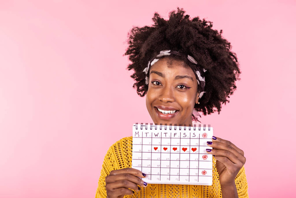 Portré mosolygós fiatal afro-amerikai nő gazdaság időszak naptár elszigetelt rózsaszín háttér. Gyönyörű fiatal nő áll elszigetelt felett rózsaszín háttér, mutatja a menstruációs naptár - Fotó, kép