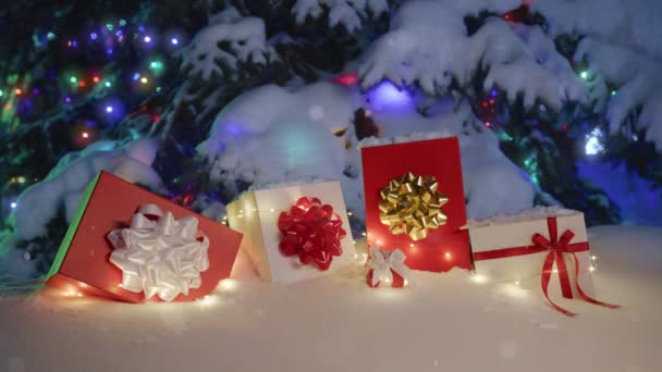 Nieve escénica en el fondo de días festivos. Muchas cajas de regalo rojas y blancas y decoraciones de Navidad bajo un hermoso árbol de Navidad nevado decorado con luces bokeh en el bosque mágico de invierno en la noche nevada - Imágenes, Vídeo