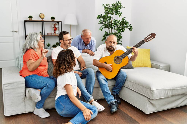 Група друзів середнього віку вечірка, яка грає на класичній гітарі, сидить на дивані вдома
. - Фото, зображення