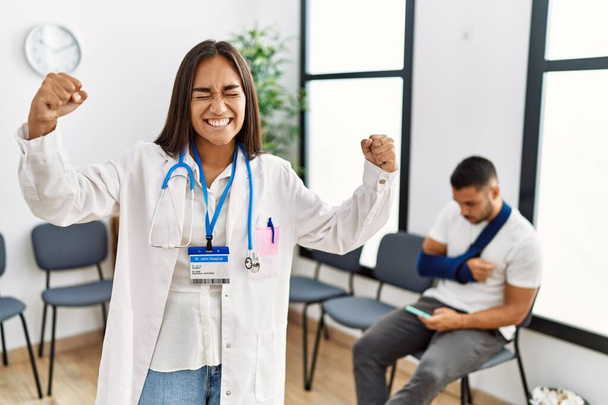 Junge asiatische Ärztin im Wartezimmer mit einem Mann mit gebrochenem Arm sehr glücklich und aufgeregt dabei Siegergeste mit erhobenen Armen, lächelnd und schreiend nach Erfolg. Festkonzept.  - Foto, Bild