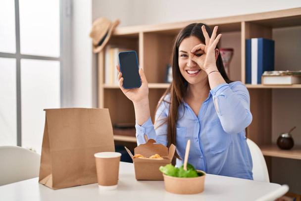 Mladá brunetka žena jíst odnést jídlo doma ukazující smartphone obrazovka s úsměvem šťastný dělá ok znamení s rukou na oku dívá skrz prsty  - Fotografie, Obrázek