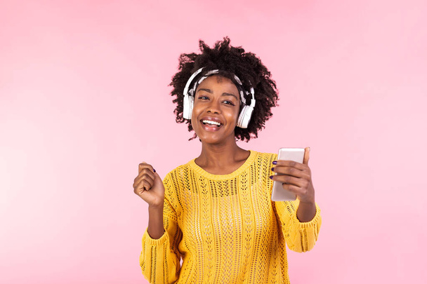 Életmód koncepció - portré gyönyörű afro-amerikai nő örömteli zenét hallgatni a mobiltelefon. trendi elegáns csinos lány fülhallgató hallgat zene tánc elszigetelt rózsaszín háttér - Fotó, kép