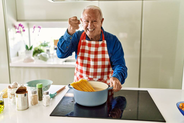 Senior man met grijs haar koken spaghetti thuis keuken geïrriteerd en gefrustreerd schreeuwen van woede, schreeuwen gek van woede en hand opgevoed  - Foto, afbeelding