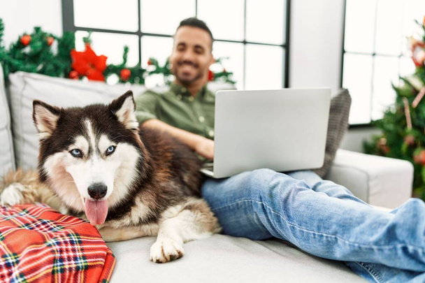 Junger hispanischer Mann sitzt mit Laptop auf Sofa mit Hund am Weihnachtsbaum - Foto, Bild