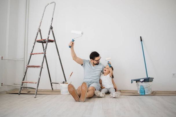 Young father and his little blonde cute girl daughter with wall paint rollers sitting on the floor during home renovation on white wall background. - Фото, зображення