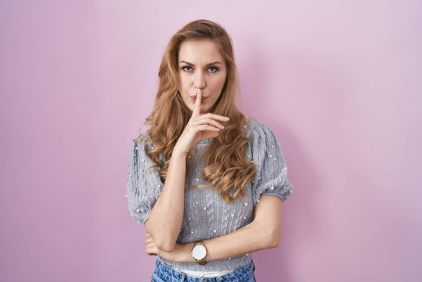 Beautiful blonde woman standing over pink background asking to be quiet with finger on lips. silence and secret concept.  - Фото, изображение