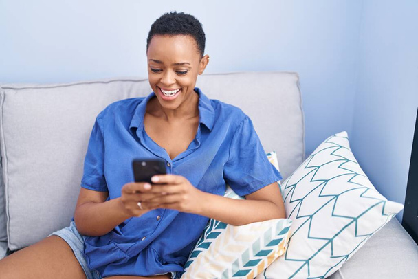 Mujer afroamericana usando teléfono inteligente sentado en el sofá en casa - Foto, Imagen