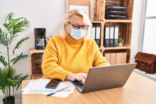 Blonde Geschäftsfrau mittleren Alters mit medizinischer Maske arbeitet im Büro - Foto, Bild