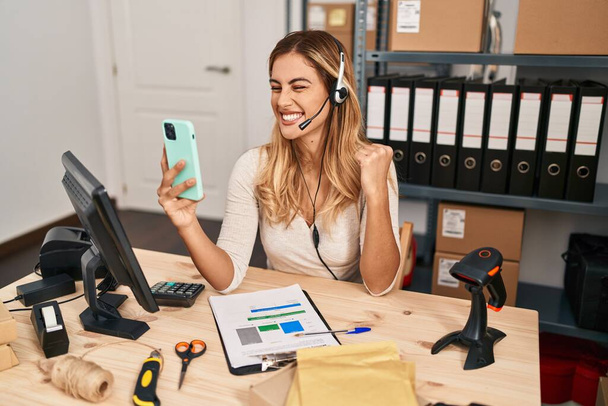 Jonge blonde vrouw werken bij kleine bedrijven e-commerce met behulp van headset schreeuwen trots, vieren overwinning en succes zeer enthousiast met opgeheven arm  - Foto, afbeelding