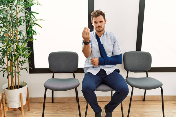 Όμορφος νεαρός άνδρας κάθεται στο γιατρό αίθουσα αναμονής με τραυματισμό στο χέρι κάνει ιταλική χειρονομία με το χέρι και τα δάχτυλα έκφραση αυτοπεποίθηση  - Φωτογραφία, εικόνα