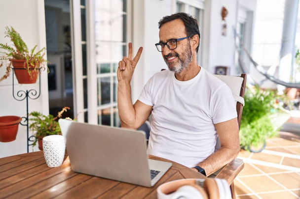 Middle age man using computer laptop at home smiling looking to the camera showing fingers doing victory sign. number two.  - Photo, image
