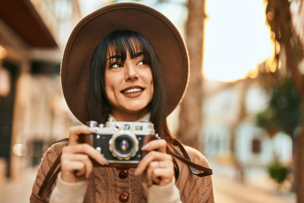 Brunetka kobieta w czapce zimowej uśmiecha się przy użyciu zabytkowej kamery na świeżym powietrzu w mieście - Zdjęcie, obraz
