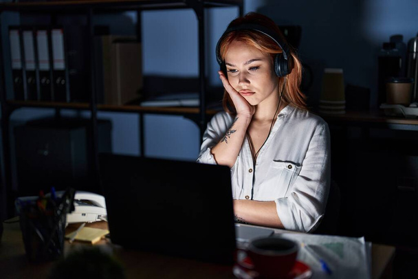 Młoda biała kobieta pracująca w biurze w nocy myśląc zmęczona i znudzona depresją z skrzyżowanymi ramionami.  - Zdjęcie, obraz